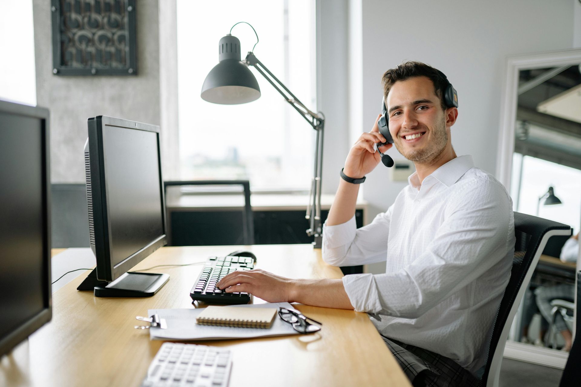 Man smiling at a desk in a bright office, talking on a phone beside a computer and lamp