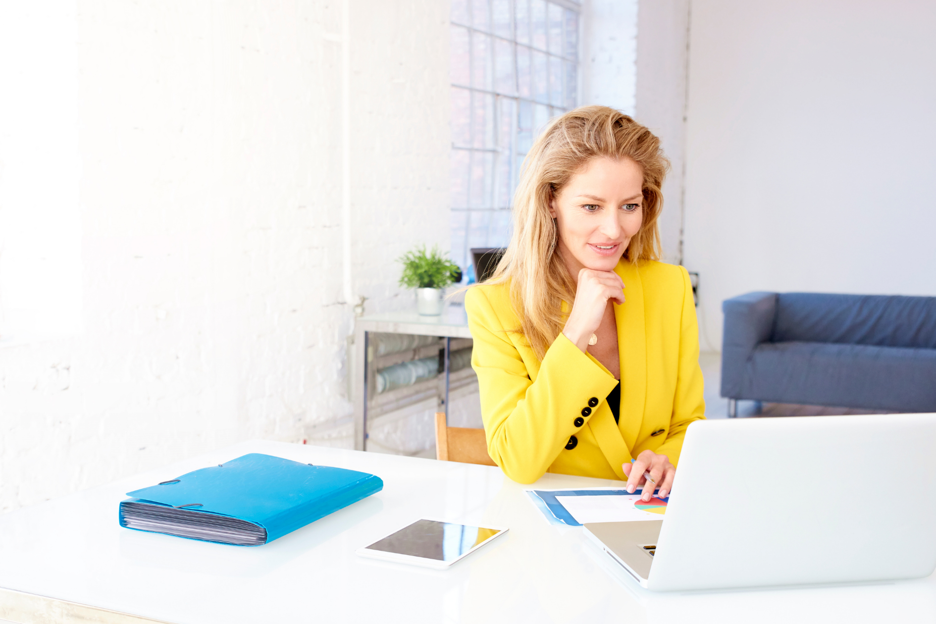 Woman in a yellow cardigan working at a desk with a laptop and notebook in a bright office
