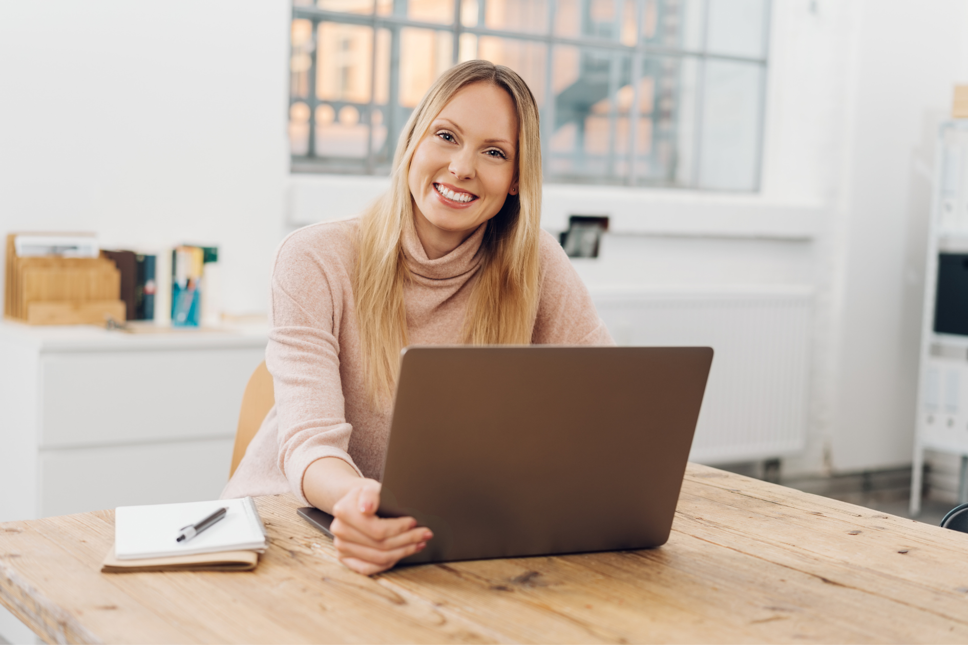 Smiling woman using a laptop at a wooden table in a bright home office