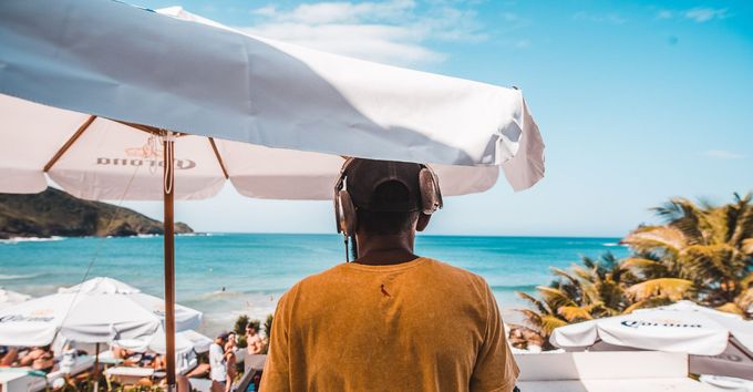 DJ usando fones de ouvido com vista para uma praia, sob guarda-sóis brancos. Dia ensolarado.