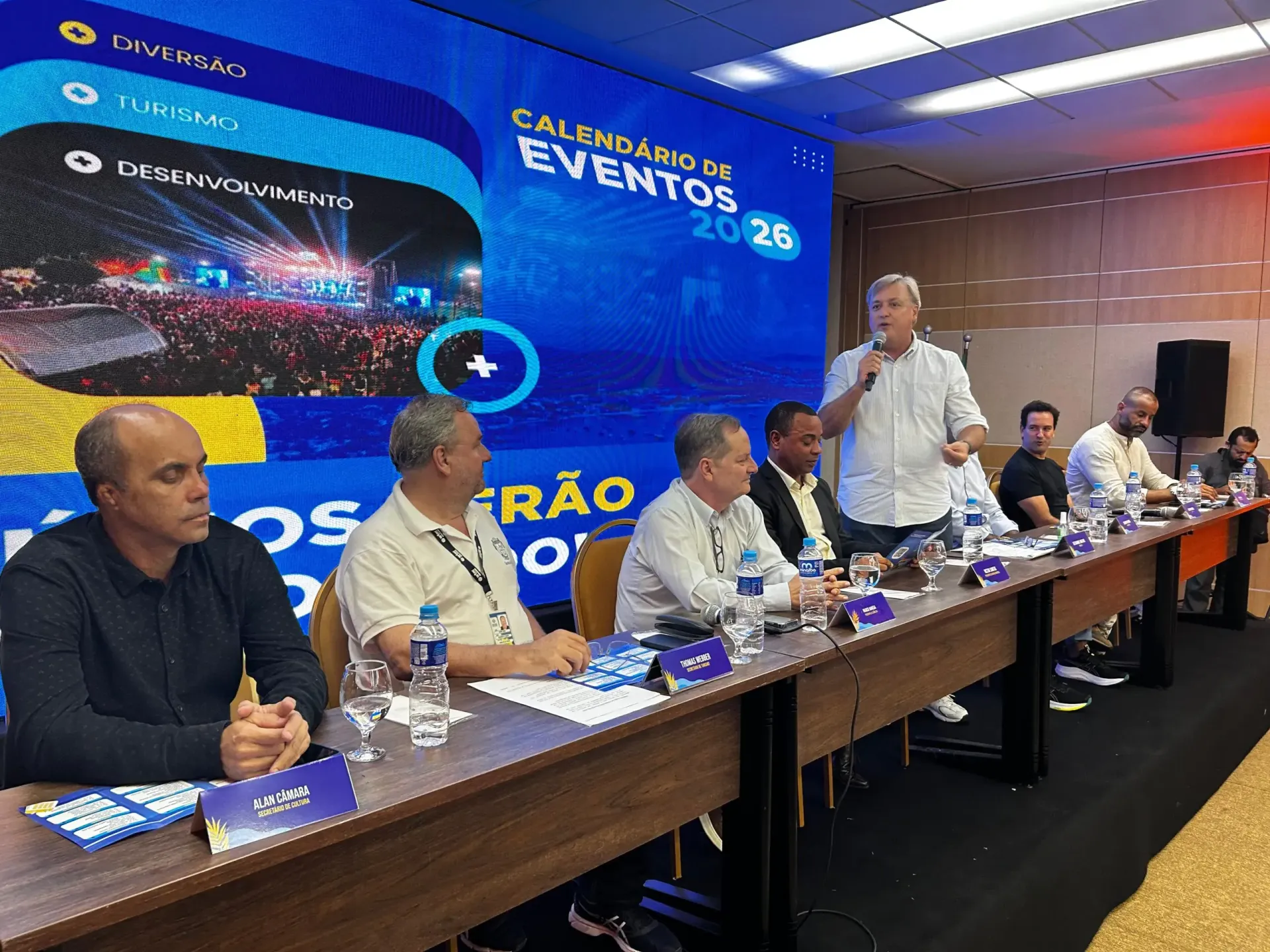 Um homem conversa à mesa com outros homens; ao fundo, vê-se uma faixa com o 