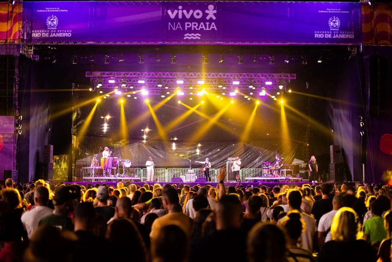 Banda se apresenta em um palco iluminado por holofotes; grande público assiste. Faixa com a inscrição 