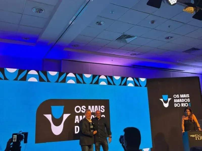 Duas pessoas em um palco com um grande telão azul exibindo 