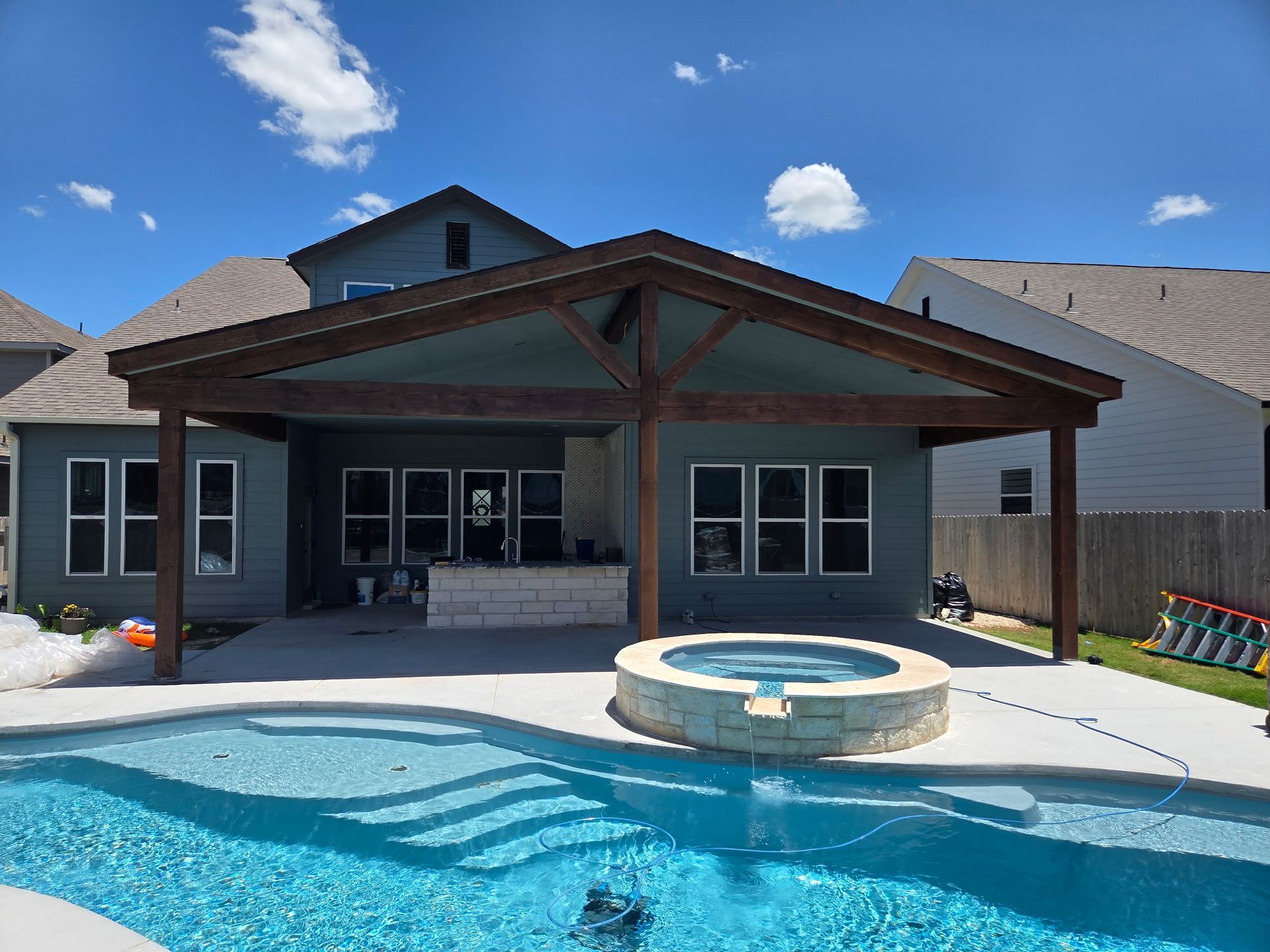 Backyard with a pool, covered patio, and house. Blue pool water, blue house with brown wood trim, sunny day.