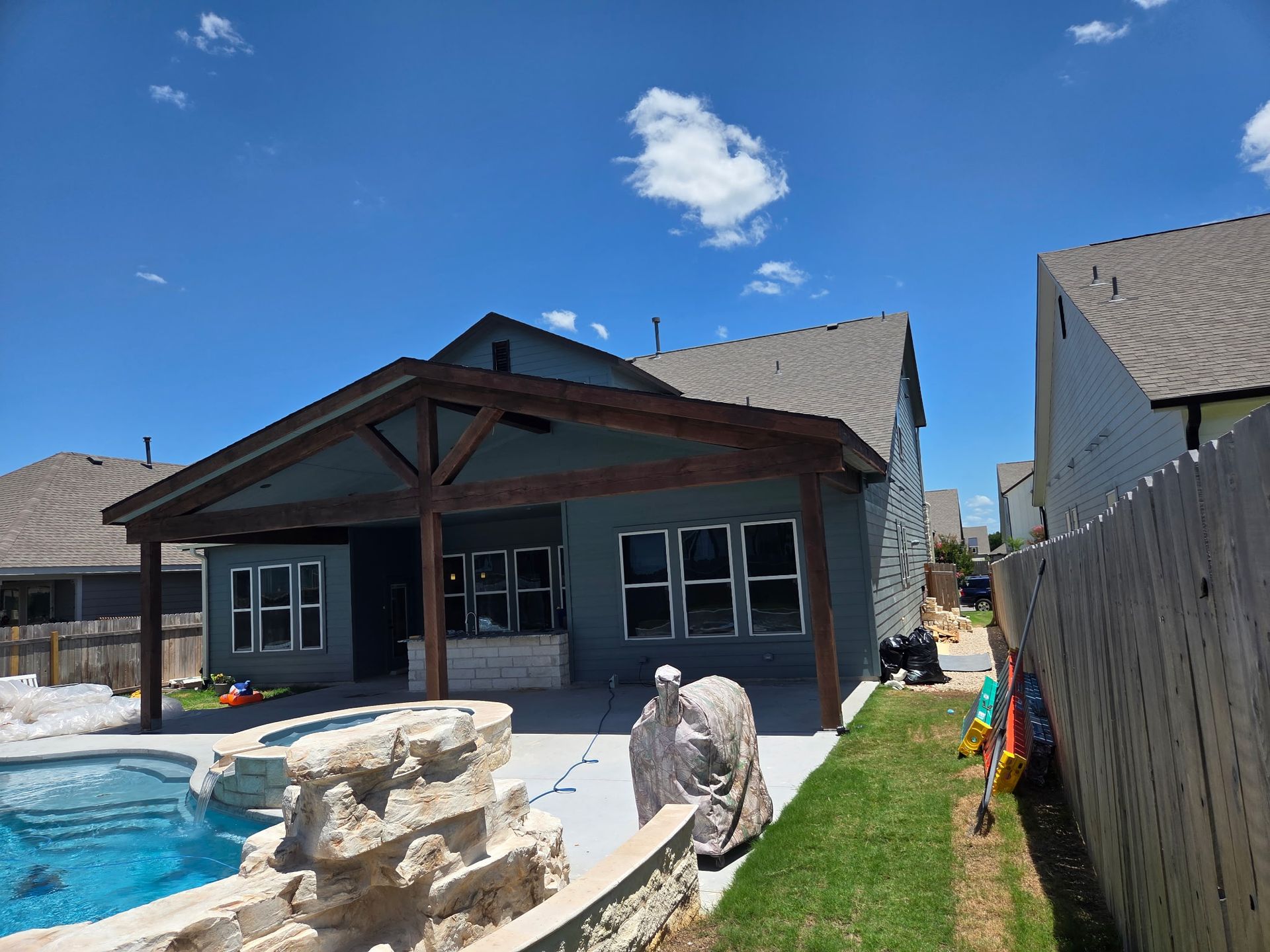Backyard with pool, patio, and covered seating area. Blue house with dark wood accents, blue sky.