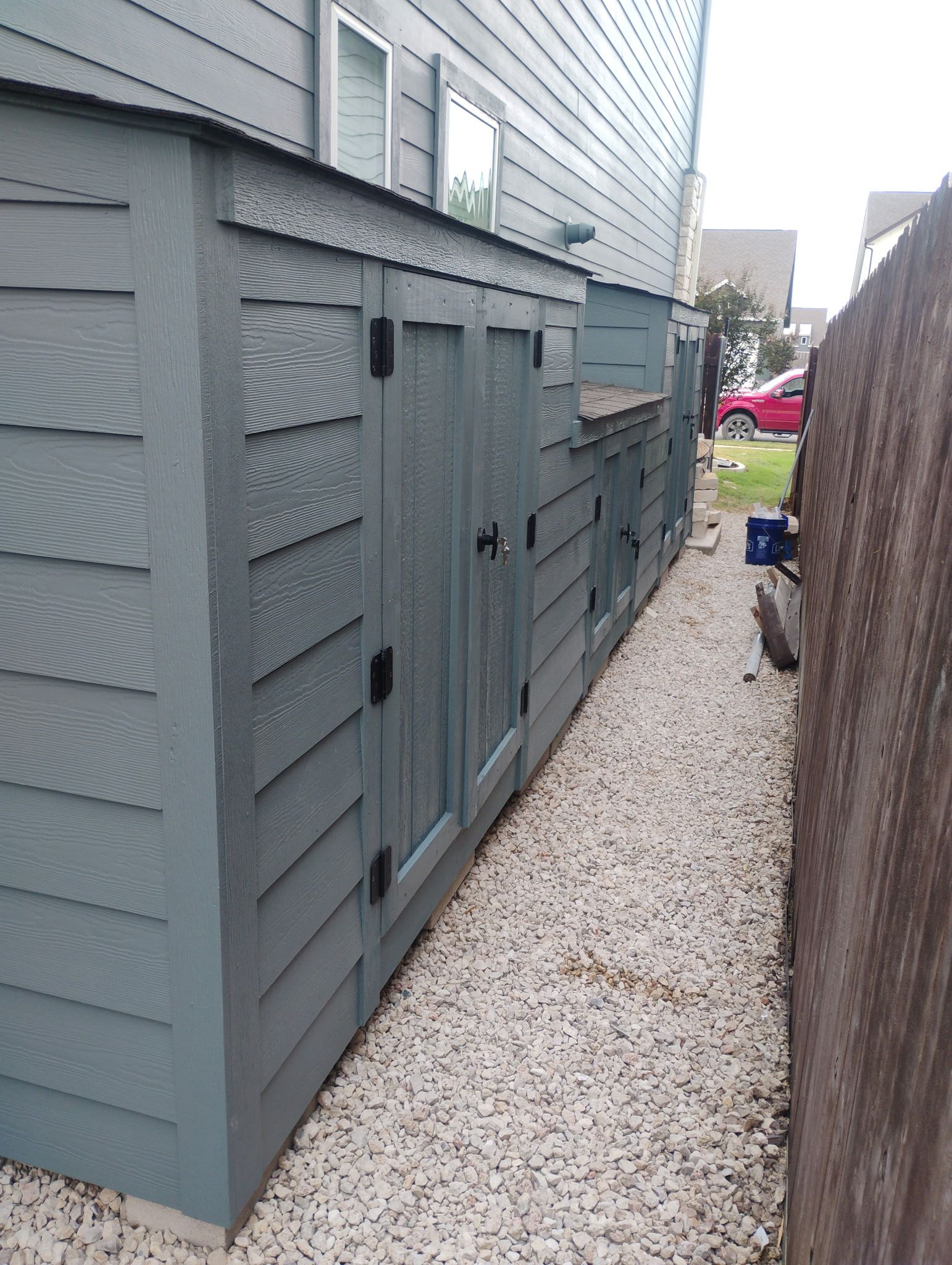 A narrow alley with blue siding, a storage unit, and a pebble pathway. A wooden fence is on the right.