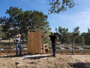 Two men standing by a wooden gate in front of a fenced garden; sunny day.