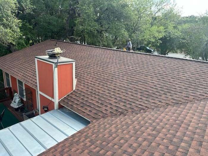 Brown shingled roof with a chimney and person working. Green trees in the background.