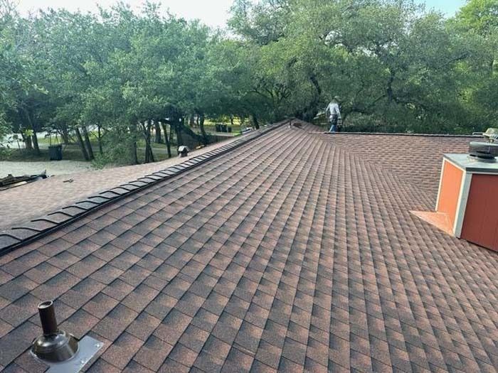Roof with brown shingles, chimney, and a person working near trees.
