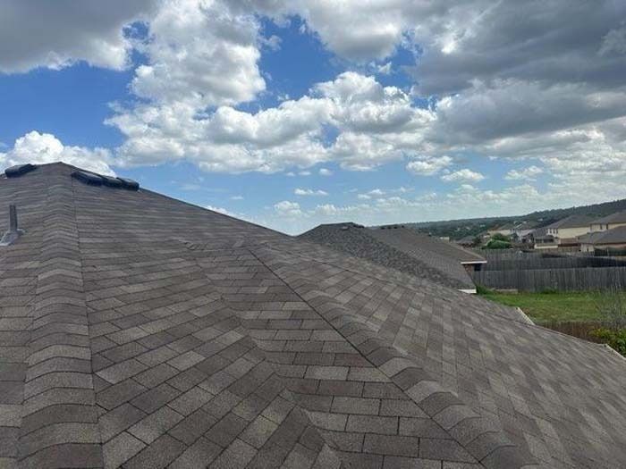 Newly shingled brown roof against a blue sky with puffy clouds; view of rooftops and distant houses.