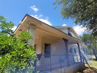 A house with a porch, tan trim, a blue sky, and green foliage.