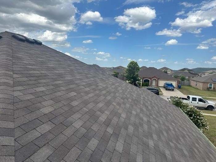 Dark gray shingle roof with blue sky, clouds, and suburban houses in background.