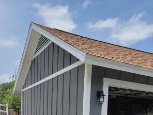 Gray shed with brown roof and white trim against a blue sky with clouds.