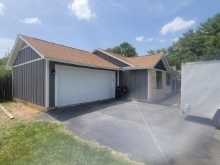 Garage with gray siding, white garage door, brown roof, and black driveway on a sunny day.