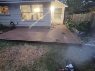 Brown deck extending from a light blue house with windows. Green grass surrounds the deck, with a fence and plants on the right.