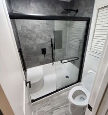 Bathroom with a black-framed shower, gray tiled walls, toilet, and a window with white blinds.