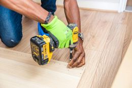 Person using a power drill to assemble wooden furniture on a light-colored wooden floor, wearing a green glove.