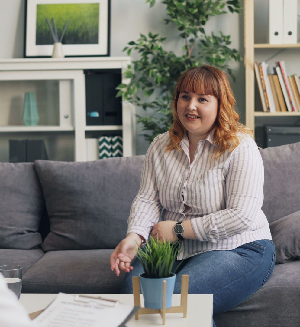 Woman sitting on a gray couch in a bright living room, smiling beside a small coffee table and plants