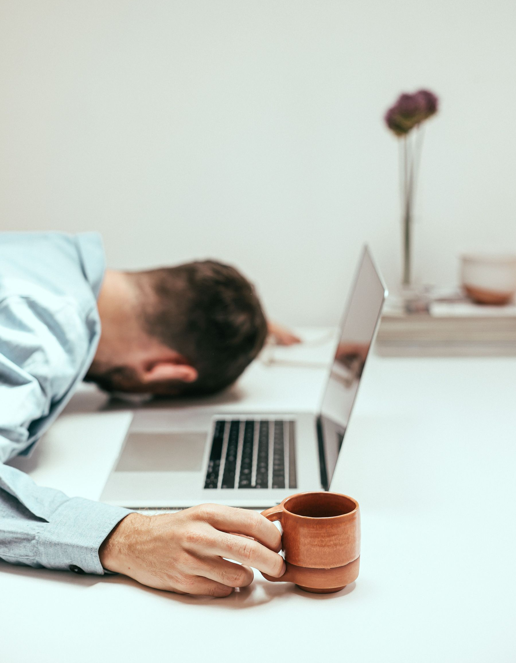 Person asleep at desk beside open laptop, hand resting on a coffee mug.