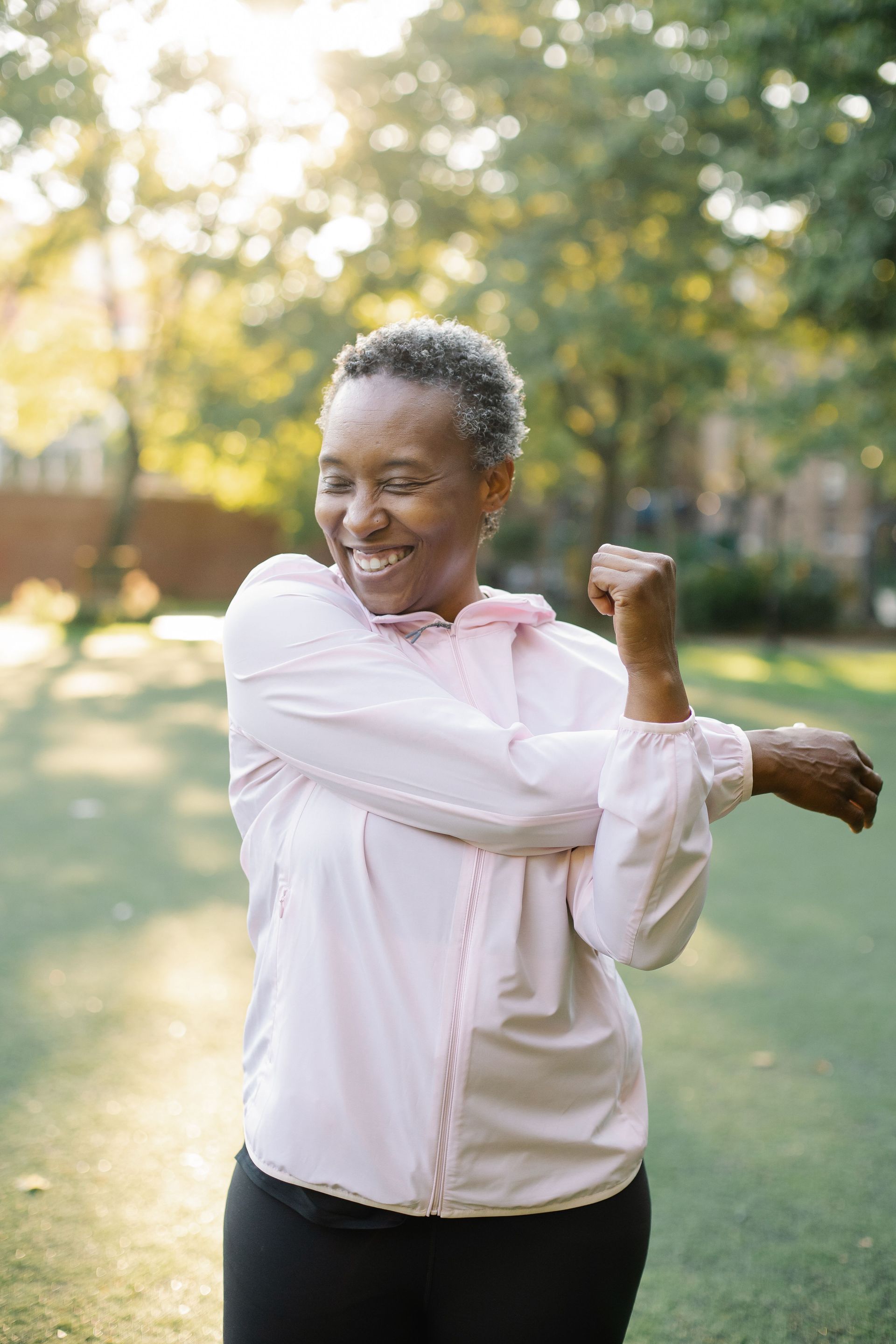 Person stretching outdoors in a park, wearing a light pink shirt and smiling in sunlight