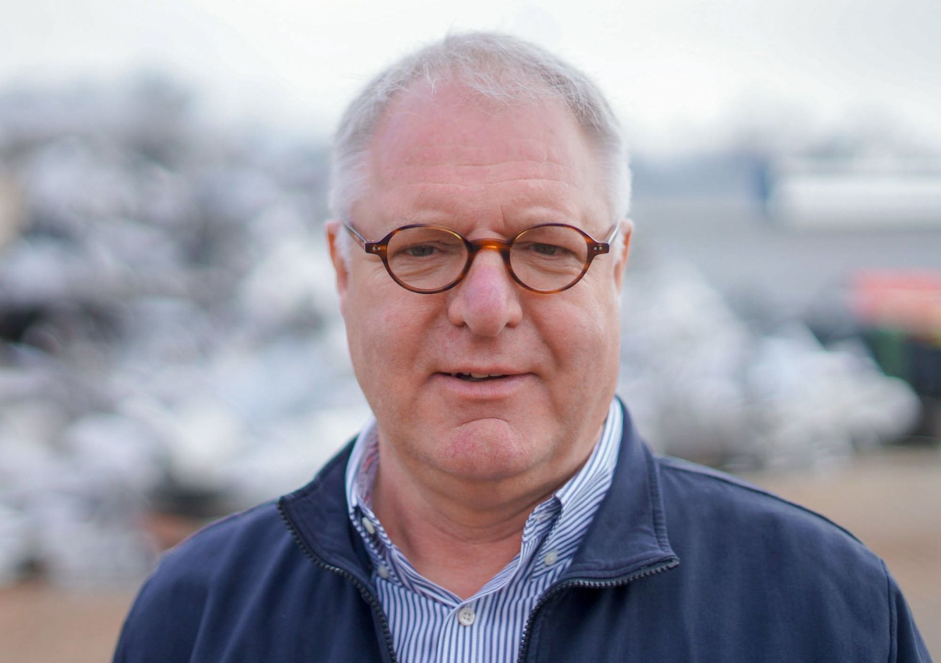Helmut Rüschen mit Brille und blauer Jacke steht vor einem Haufen Autos.