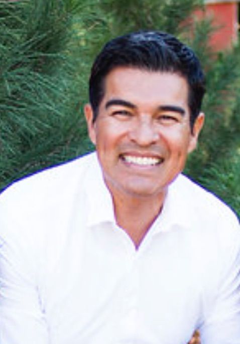 Man with dark hair smiling, wearing a white button-up shirt; outdoors with green foliage.