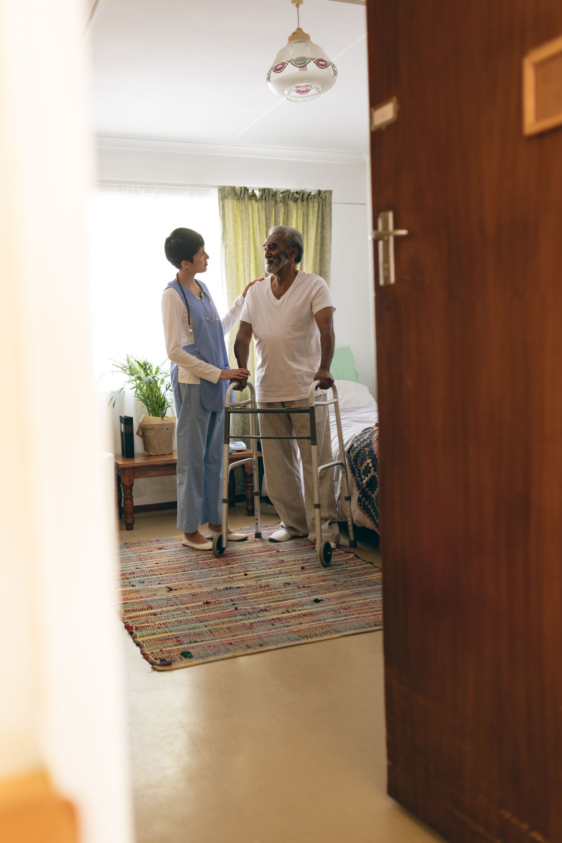 Caregiver assists a person using a walker in a room with a patterned rug and open door.
