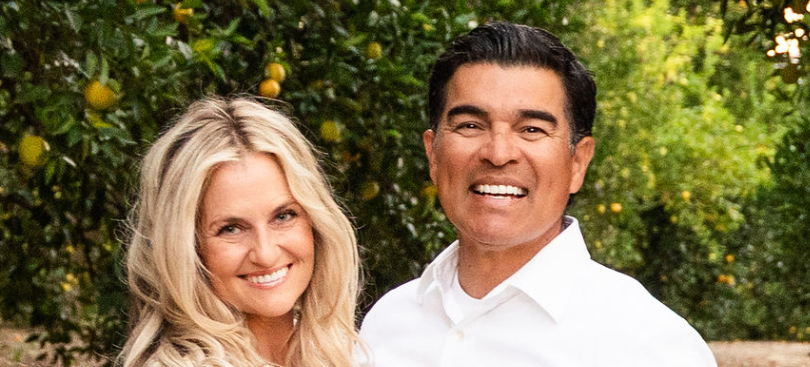 A smiling woman and man stand in front of a tree. They are wearing white shirts.