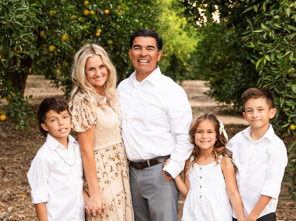 Family of five smiles in an orchard. The adults are flanked by three children, all wearing white.