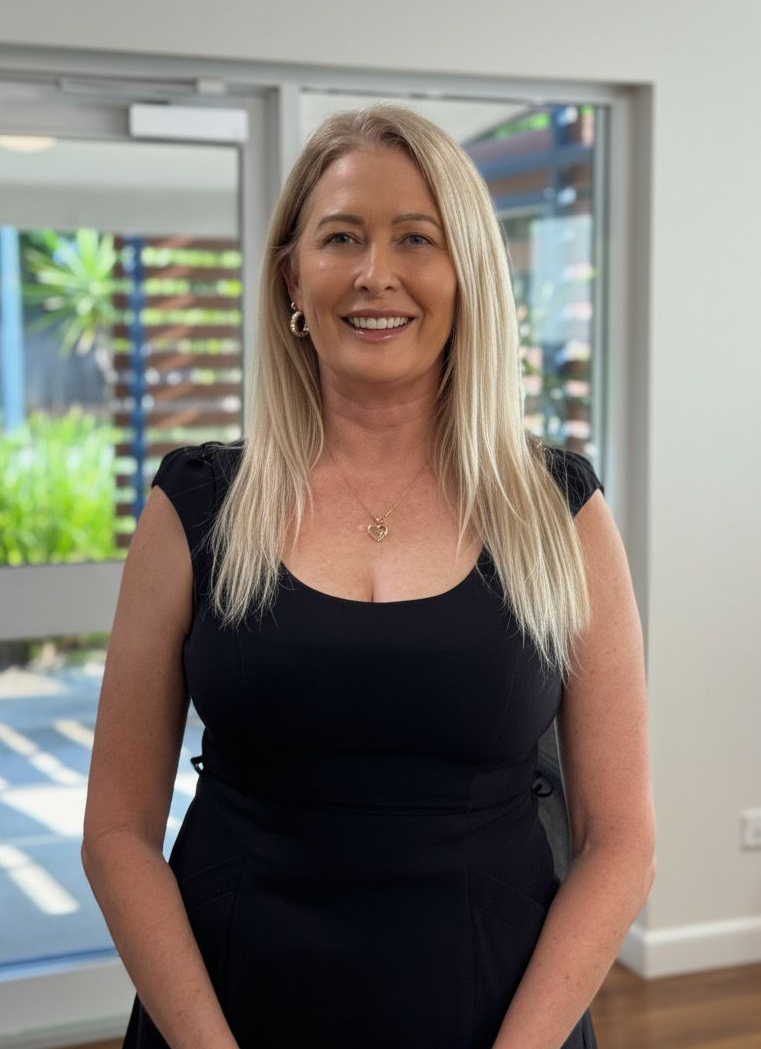 Woman with blonde hair smiling, wearing a black dress, standing indoors.