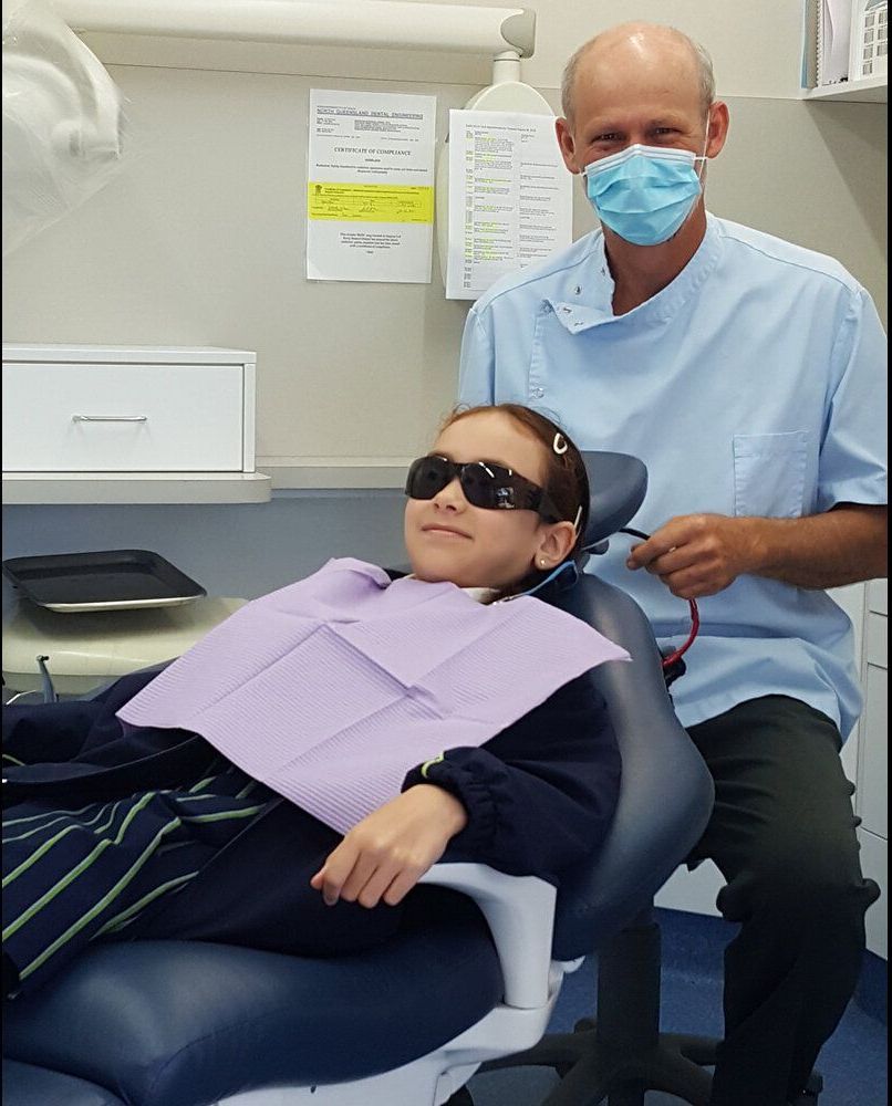 A Man Wearing A Mask Is Sitting Next To A Child In A Dental Chair — Barry Bennett Dental In Cairns, QLD