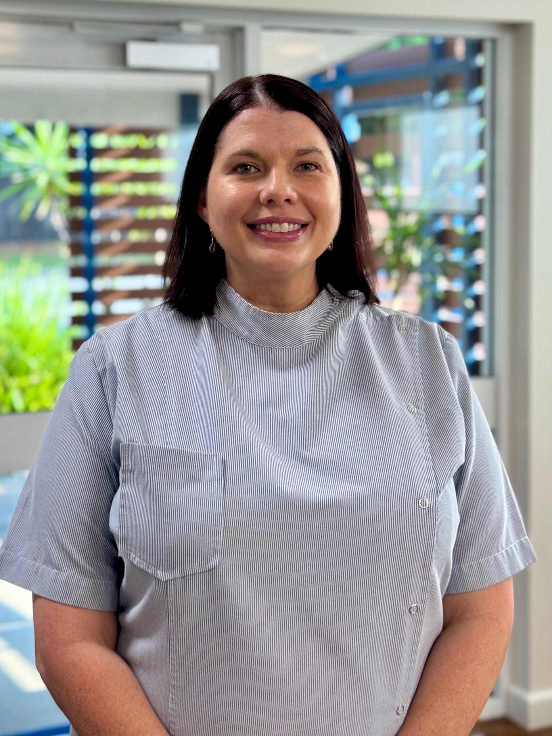 Woman with dark hair, smiling, wearing a striped shirt, standing near a window.
