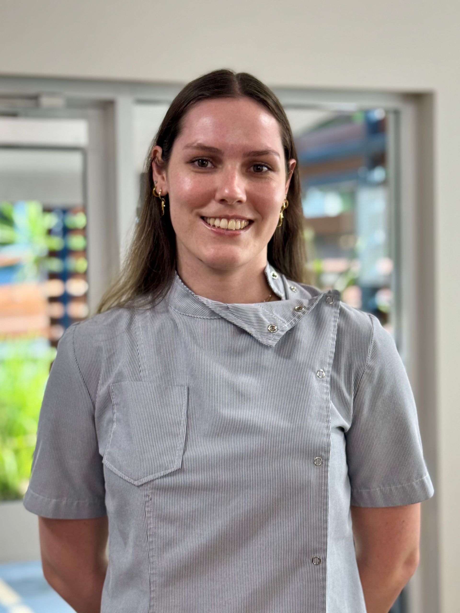 Woman with long brown hair smiling, wearing a gray striped collared shirt. Standing indoors.