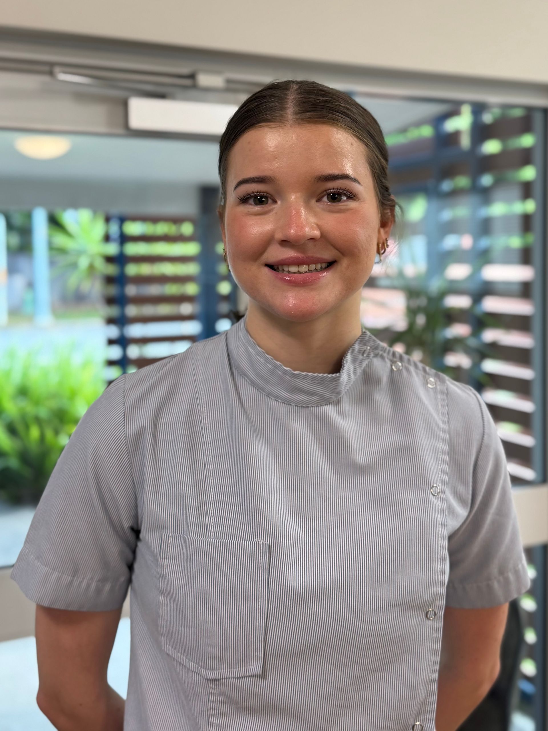 Woman smiling, wearing a striped uniform, standing indoors with a glass door in the background.