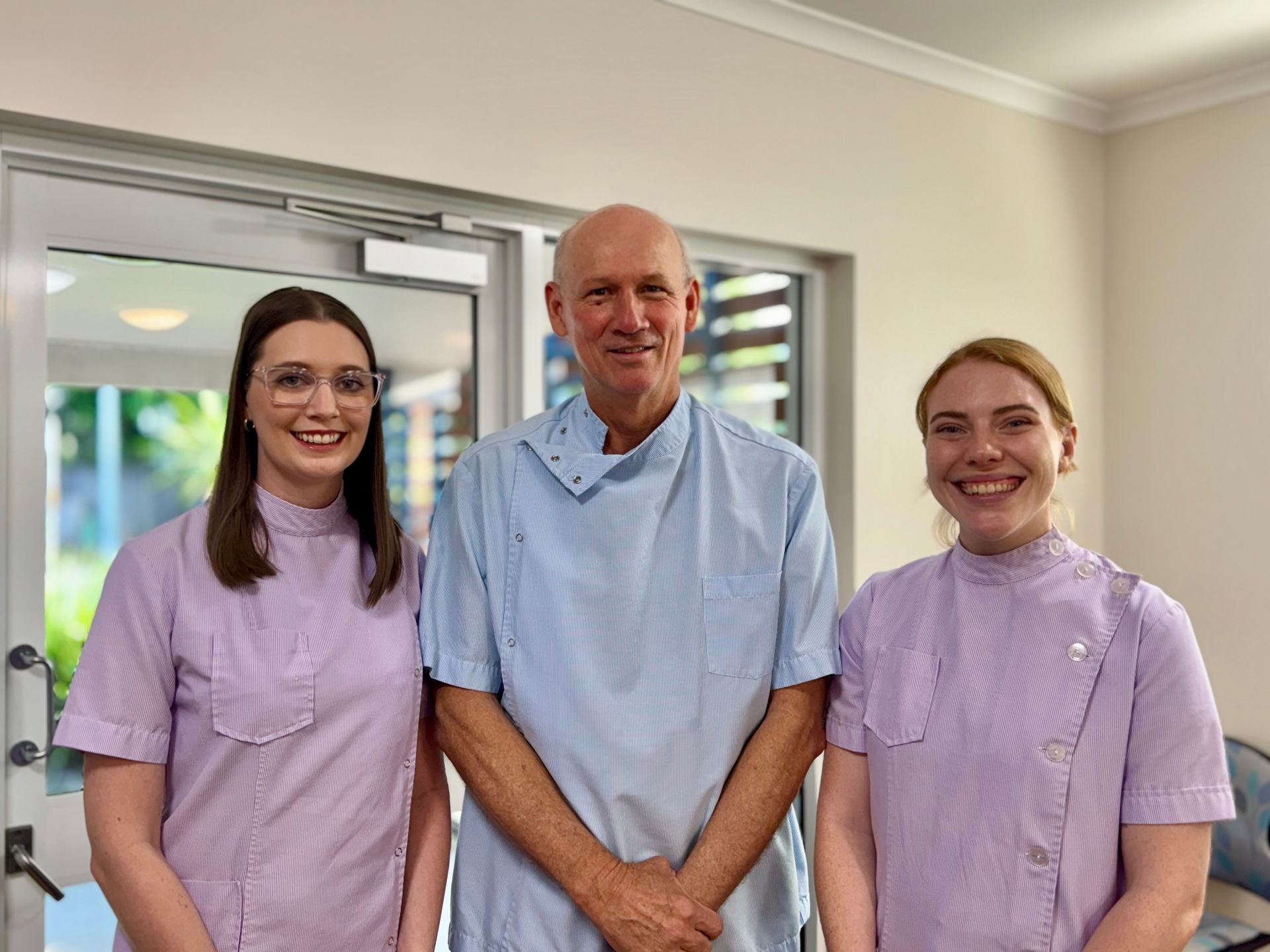 Three dental professionals standing indoors; two in lilac scrubs flanking a man in a blue scrub top, smiling.