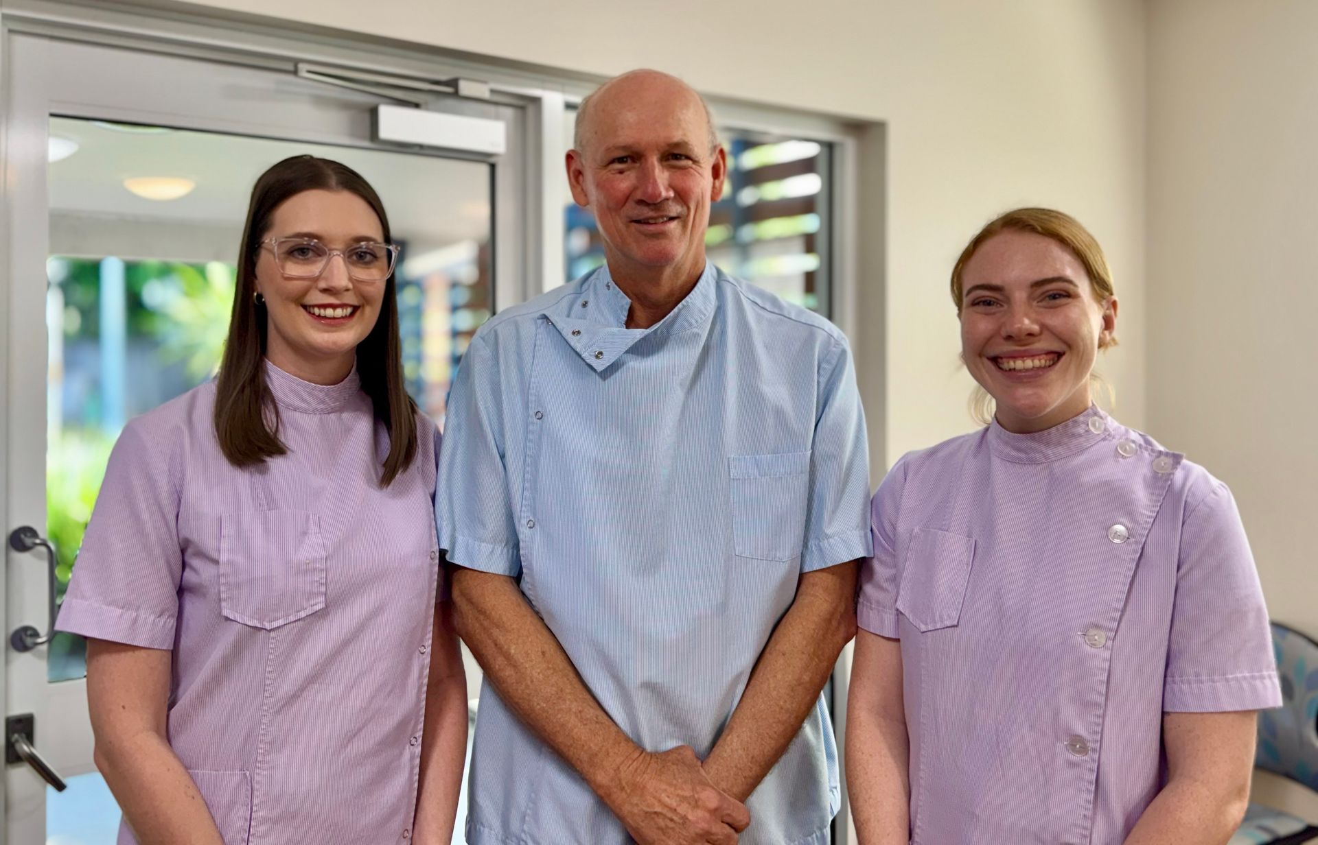 Three healthcare workers smiling in a clinic. Two women wear purple uniforms, one man in a blue scrub top.