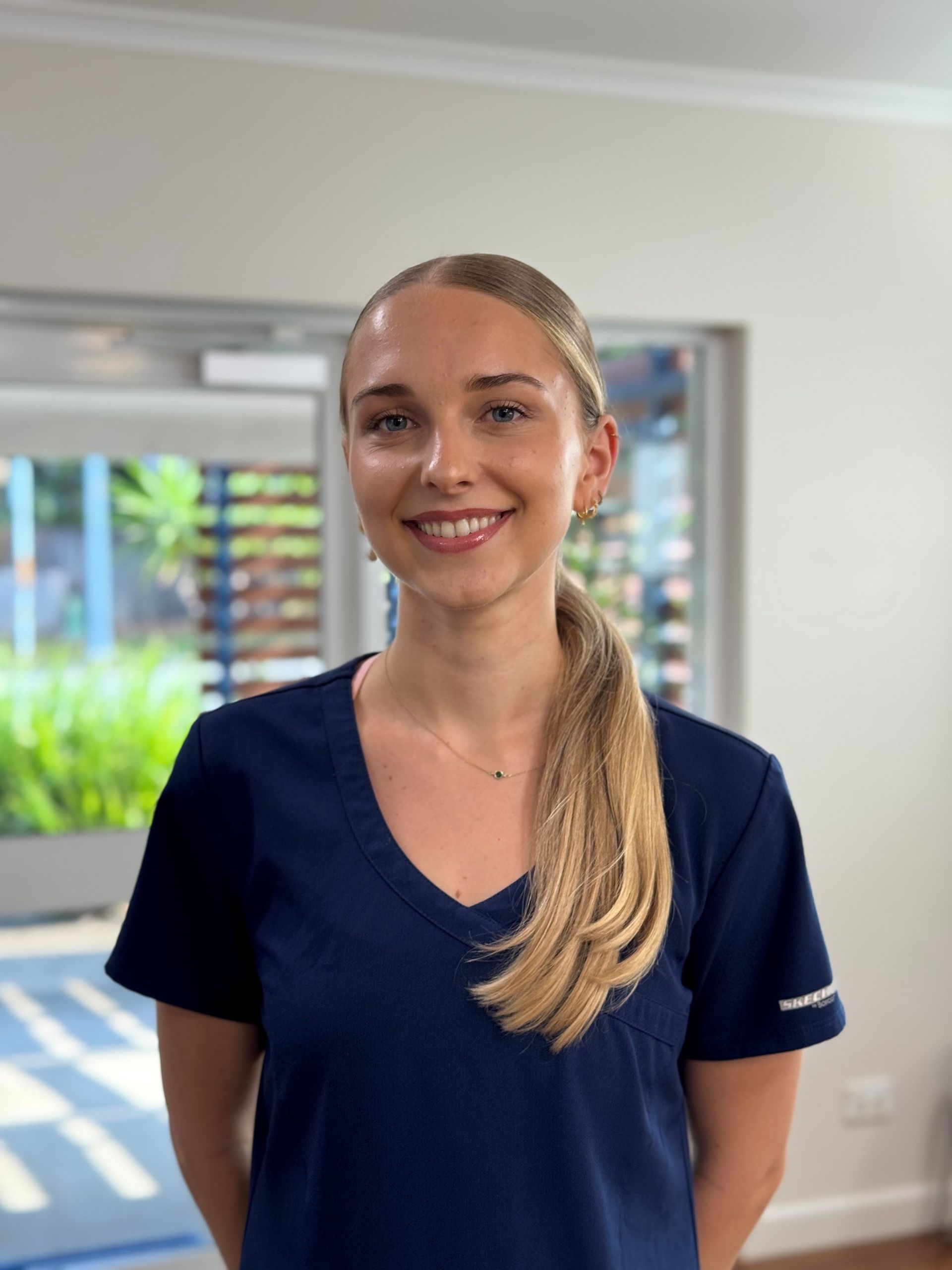 Woman with blonde ponytail smiling, wearing a navy blue v-neck shirt, standing indoors.