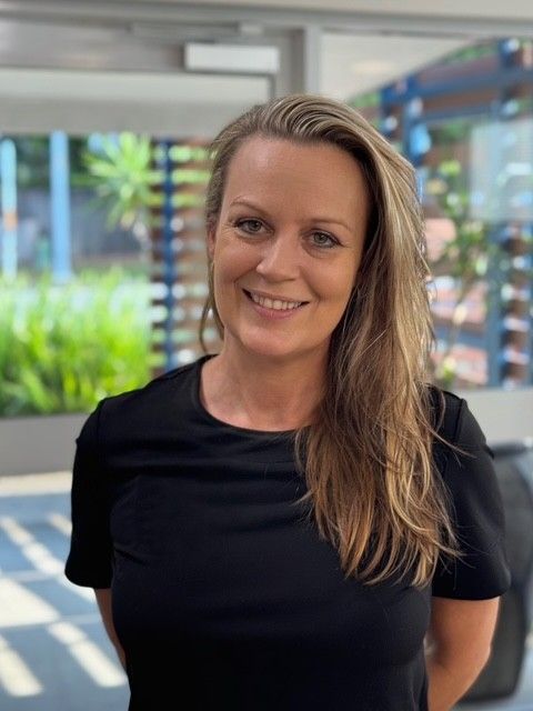Woman with blonde hair smiles at the camera, wearing a black shirt in a bright hallway.