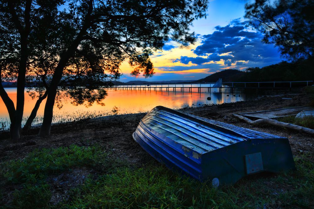 A boat sits on the shore of a lake at sunset — Bay Blinds & Doors in Charmhaven, NSW