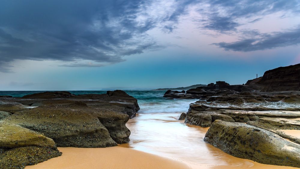 A beach with rocks and waves coming in on a cloudy day — Bay Blinds & Doors in Wamberal, NSW