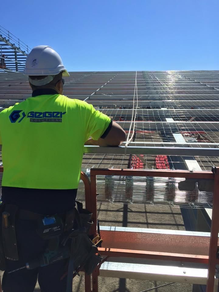 A Man Wearing a Hard Hat and a Neon Yellow Shirt is Looking at a Row of Solar Panels — Geiger Solutions In Emerald, QLD