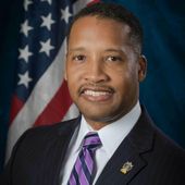 joshua bulter in a suit and tie is smiling in front of an american flag