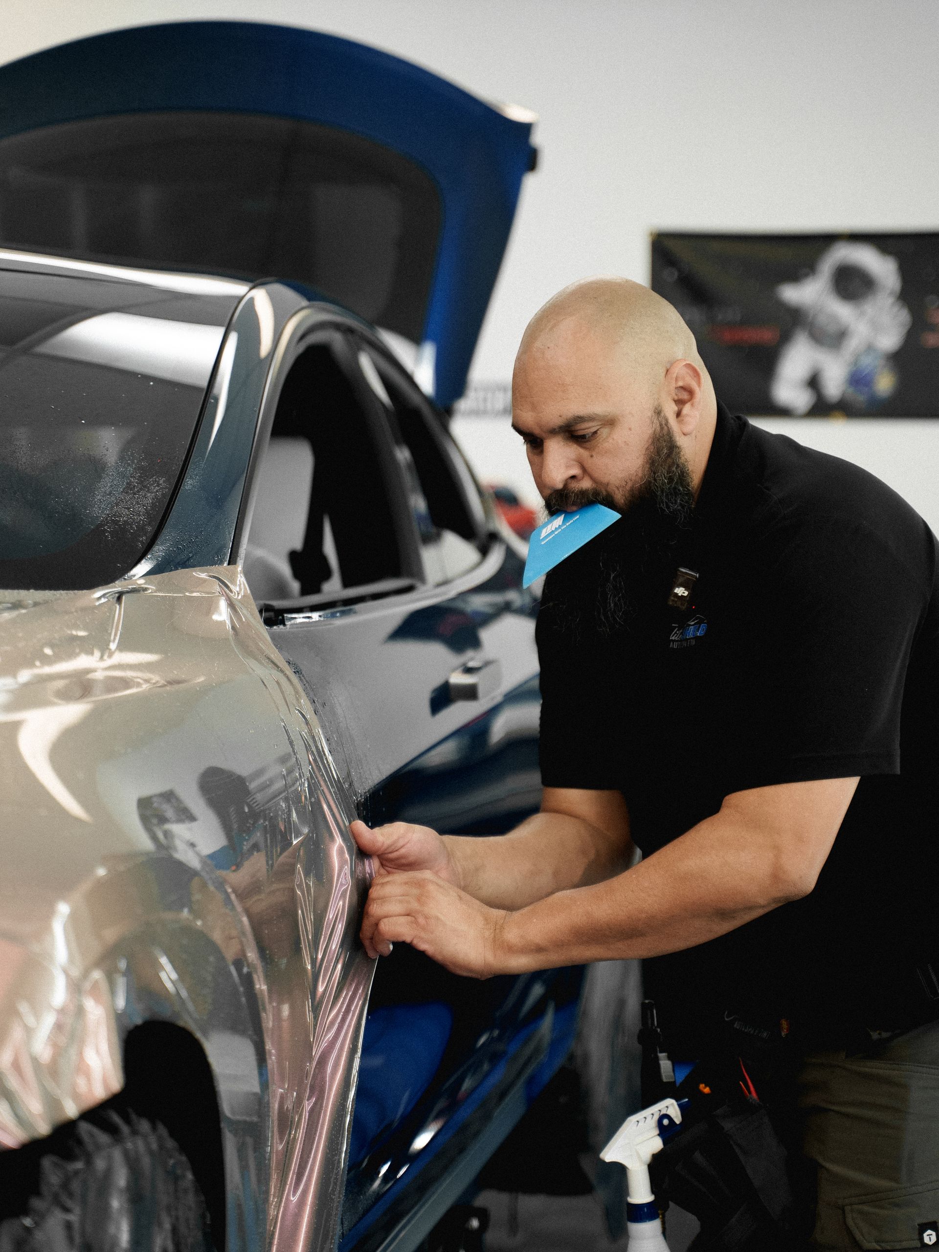 Man applying vinyl wrap to a car door. He's in a shop with a blue car, holding wrap near the door.