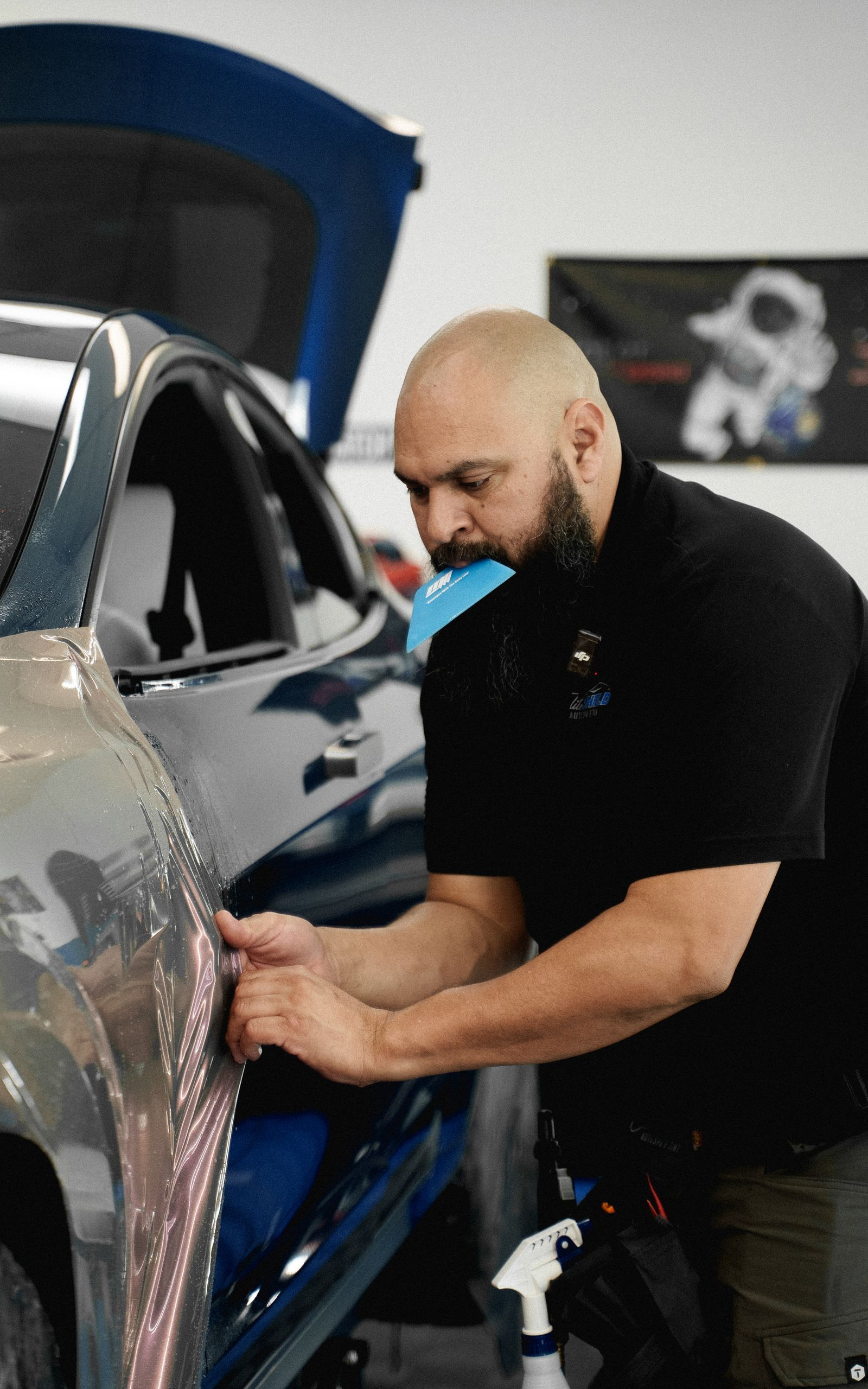 Man applying film to car door in shop. Car is blue and silver.