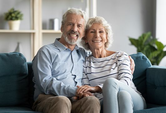 An elderly couple is sitting on a couch holding hands and smiling for the camera.