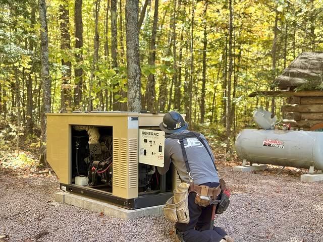 A technician in a grey uniform kneels while working on an open beige industrial generator outdoors in a wooded area.