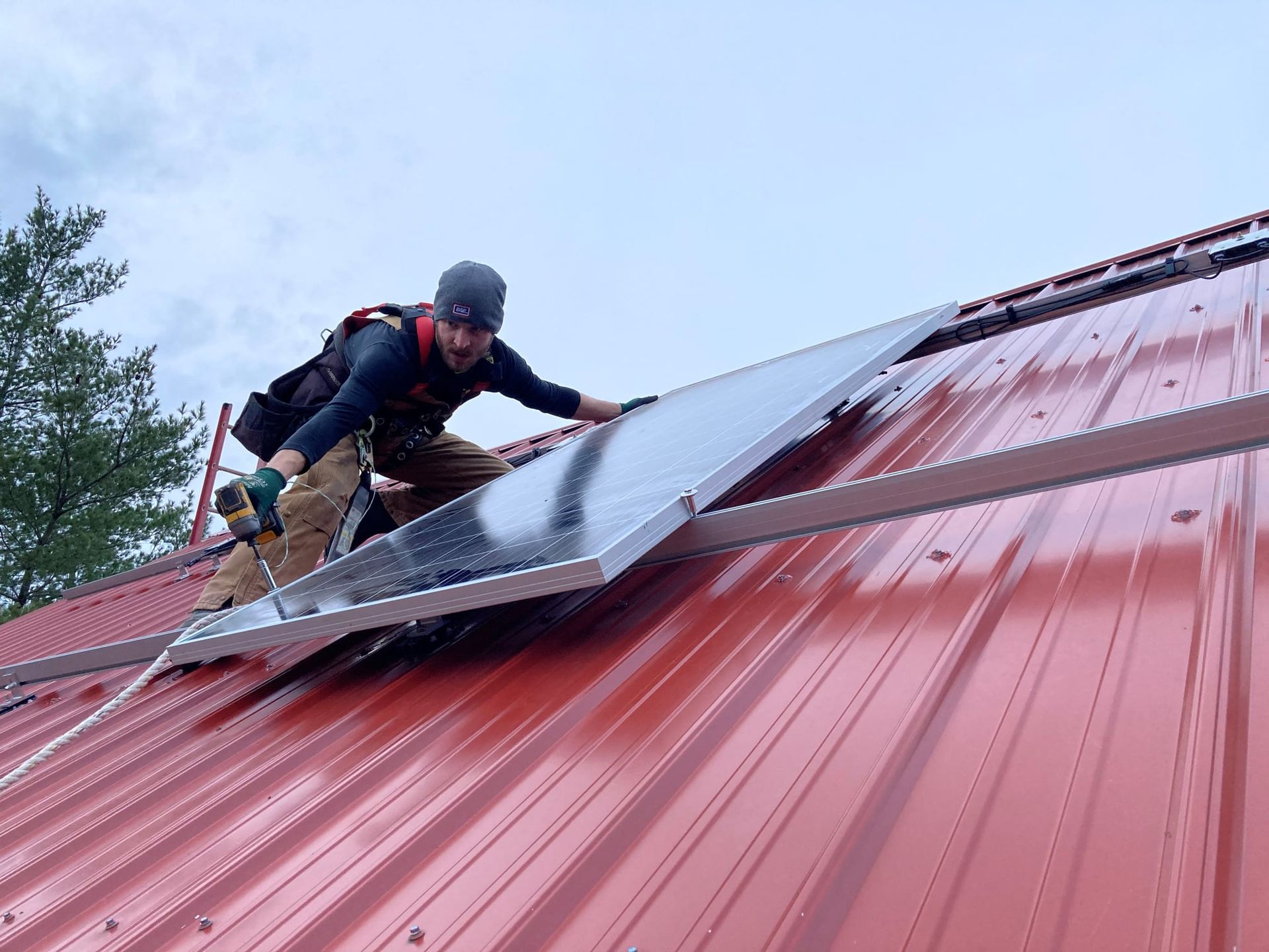 A person in a beanie and work harness installs a solar panel onto a bright red metal roof against a cloudy sky.