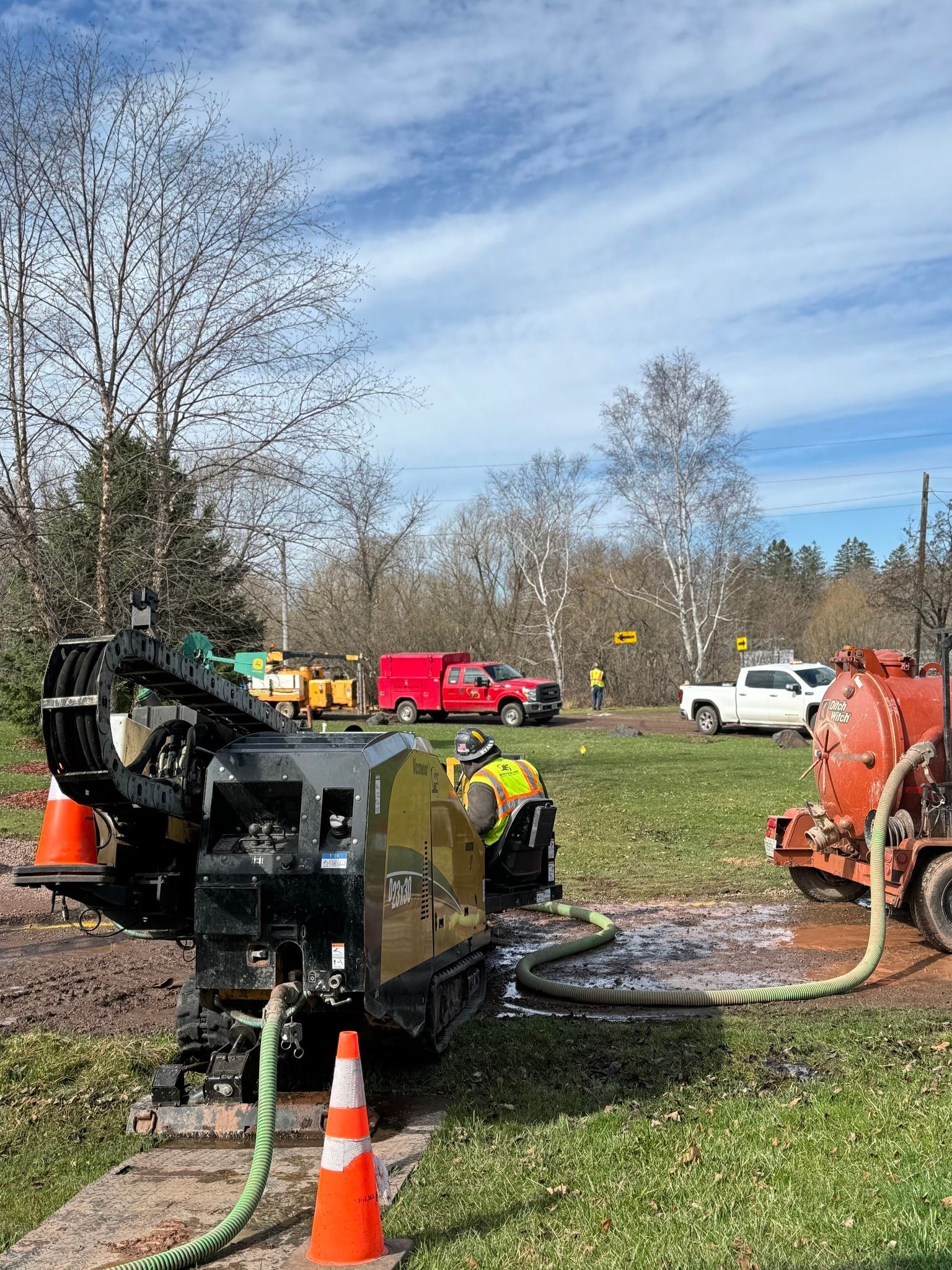 A construction crew operates a yellow horizontal directional drill in a grassy field with utility vehicles parked nearby.