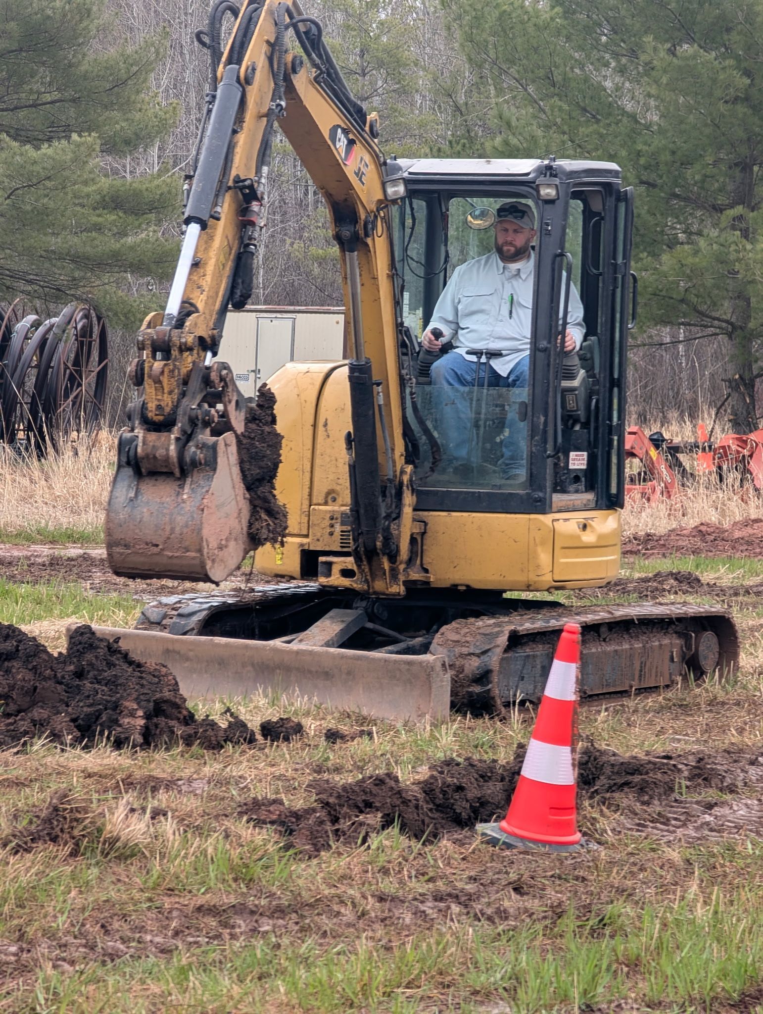 A yellow excavator with a person in the cab sits in a field near a red and white traffic cone.