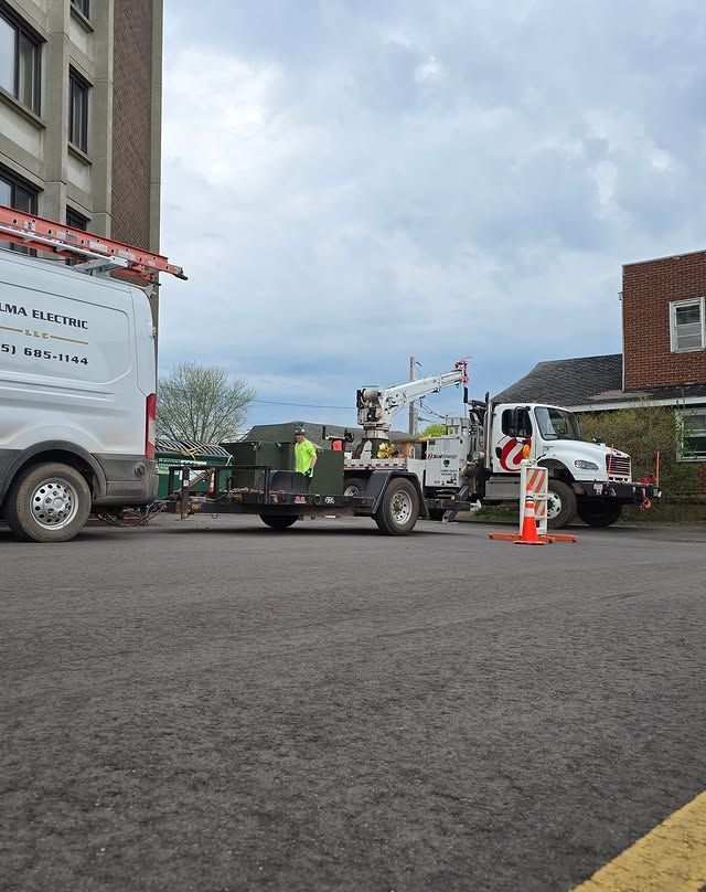 Utility truck and white van parked on an asphalt street with a worker near equipment on a flatbed trailer.